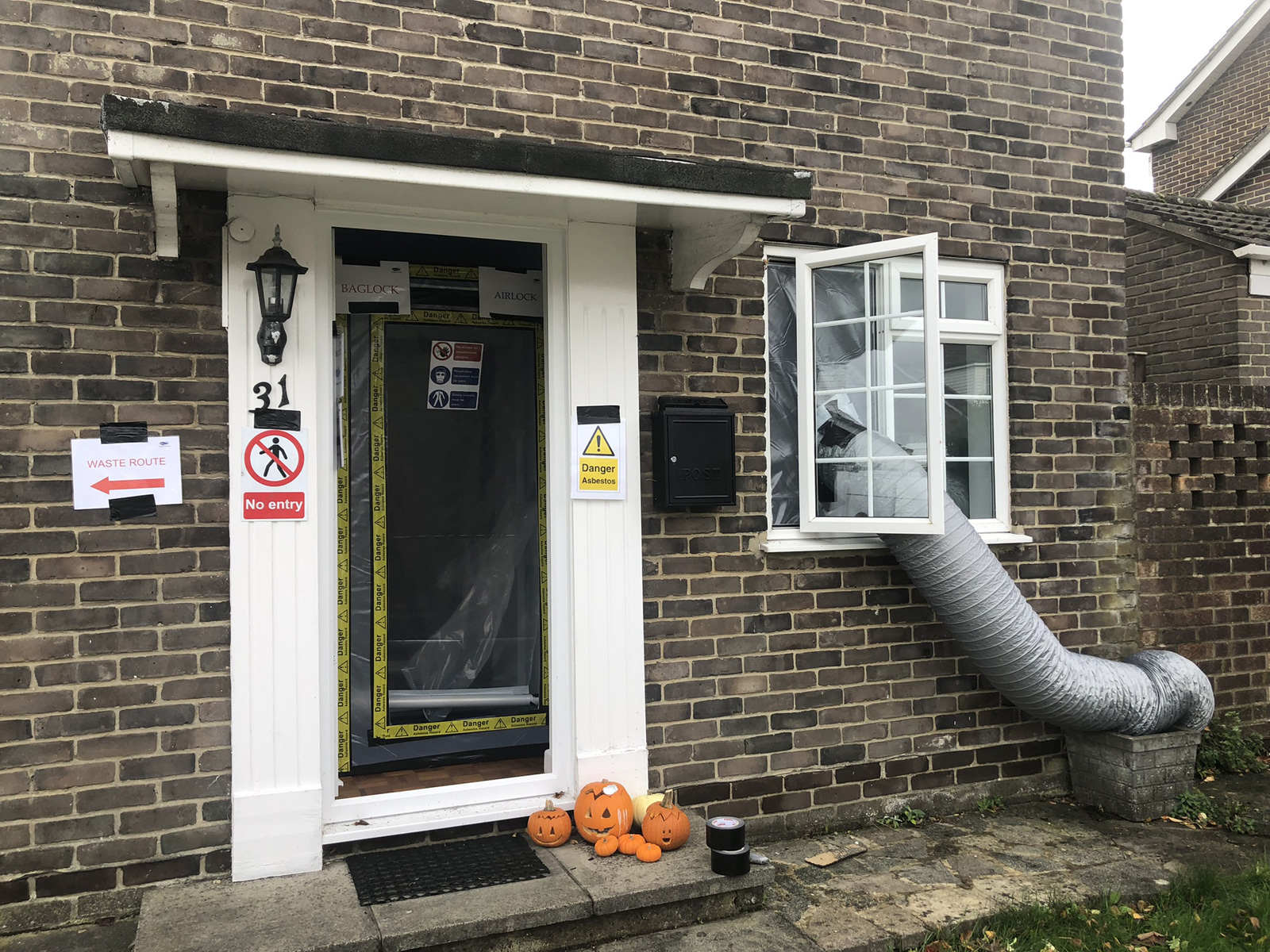 The exterior showing Halloween pumpkins on doorstep next to hazmat containment - all the professional signage (WASTE ROUTE, BAGLOCK, AIRLOCK, Danger Asbestos), grey ducting for ventilation and testing, life goes on